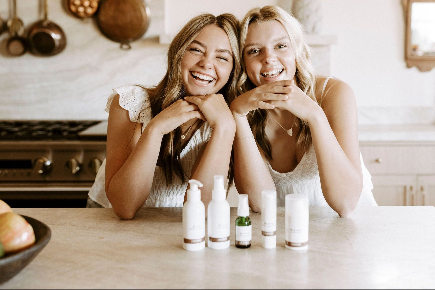 Two women sitting at a kitchen table with skincare products and fruits.